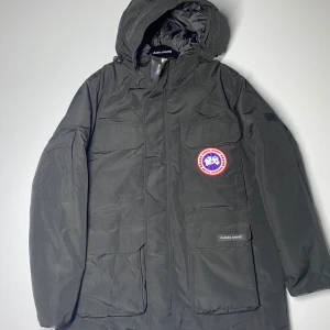 Svart parkajacka från Canada Goose - Säljer en svart parkajacka från Canada Goose i mycket bra skick. Jackan är perfekt för kalla vinterdagar med sin varma och tåliga design. Den har en huva och flera praktiska fickor. Märkets logga syns tydligt på bröstet och insidan. Perfekt för dig som vill hålla stilen i vinter! ❄️