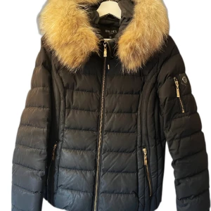 Svart dunjacka med päls från Hollies - Säljer en snygg svart dunjacka från Hollies med fluffig päls på kragen. Jackan har dragkedja och flera praktiska fickor med dragkedjor. Perfekt för kalla vinterdagar och håller dig varm och stilren. Passar både till vardags och festliga tillfällen. Nypris 3499:-💕