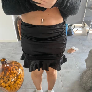 Svart sammetskjol med volang - Säljer en superfin svart sammetskjol med volangdetalj längst ner. Kjolen är tight och kort, perfekt för festliga tillfällen. Den har en elegant glans och sitter snyggt på kroppen. Passar perfekt till en mysig tröja eller en glittrig topp för en utekväll! 🖤