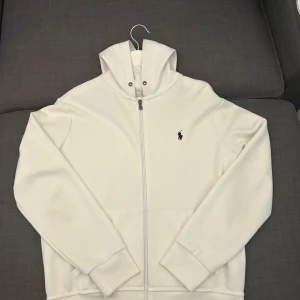 Ralph Lauren Zip Vit - Säljer en stilren vit zip hoodie från Ralph Lauren i fint skick. Den har en dragkedja framtill och den klassiska loggan broderad på bröstet. Perfekt för både höst och vår. Storlek M. Kan mötas i Stockholm. Annars frakt som gäller. Hör av dig vid funderingar.