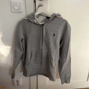 Grå hoodie från Polo Ralph Lauren - Säljer en snygg och bekväm grå hoodie från Polo Ralph Lauren. Den har en dragkedja framtill och en klassisk broderad logga på bröstet. Perfekt för både höst och vår, med långa ärmar och en skön huva. Passar till alla tillfällen, från chill till casual outings.
