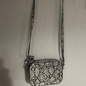 Ormmönstrad axelväska - Säljer en supercool axelväska med ormmönster. Den är perfekt för att ge din outfit en edgy touch. Väskan har en justerbar rem och dragkedja upptill. Den är i mycket bra skick och passar perfekt för både vardag och fest. Aldrig använd.
