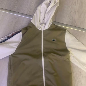 Beige och vit jacka från Lacoste - Säljer en snygg beige och vit jacka från Lacoste. Den har en dragkedja framtill och en huva. Ärmarna är vita med ett coolt mönster i flera färger längs sidorna. Perfekt för vår och höst! 🐊 Pris kan diskuteras 