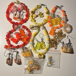 19 Färgglada smycken med pärlor och berlocker - Ett set med färgglada smycken, inklusive armband och örhängen. Armbanden har pärlor i rött, gult och orange med söta berlocker som fjärilar och björnar. Örhängena har små påsar med apelsinmotiv och björnberlocker. Perfekt för att liva upp vilken outfit som helst!