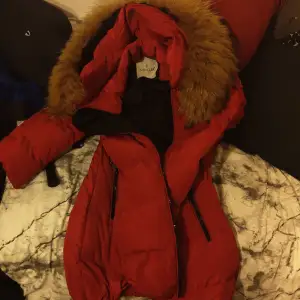 Säljer en snygg röd dunjacka från Moncler med en fluffig pälsdetalj på huvan. Jackan har en dragkedja framtill och är perfekt för kalla vinterdagar. Den är i mycket bra skick och håller dig varm och stilren hela säsongen. Passar perfekt för både vardag och festliga tillfällen!