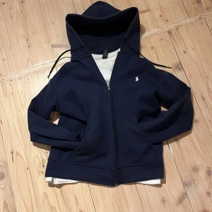 Mörkblå hoodie från Ralph Lauren - Säljer en snygg mörkblå hoodie från Ralph Lauren. Den har en dragkedja framtill och en liten broderad logga på bröstet. Perfekt för kyliga dagar och passar till allt! 