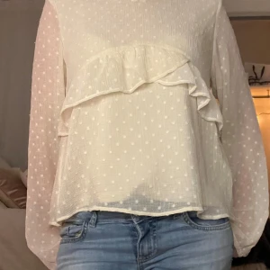 Vit blus med volangdetaljer - Säljer en söt vit blus med prickigt mönster och volangdetaljer framtill. Blusen har långa ärmar och en luftig passform, perfekt för vår och sommar. Den är gjord i ett lätt och bekvämt material, vilket gör den idealisk för både vardag och festliga tillfällen. Använd endast ett fåtal gånger