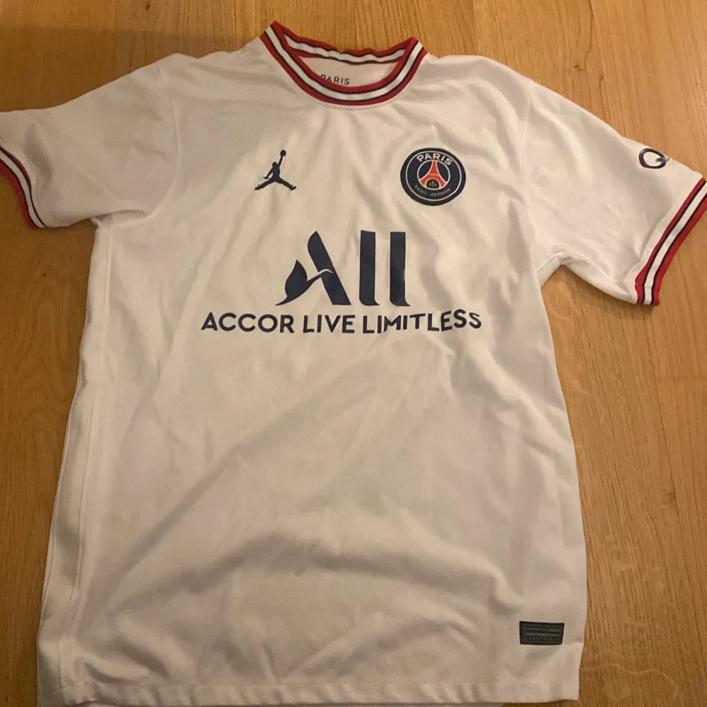 Säljer en snygg vit PSG-tröja i samarbete med Jordan. Den har korta ärmar och coola röda detaljer vid krage och ärmslut. Perfekt för alla Paris Saint-Germain-fans! Tröjan är i perfekt skick och redo för nästa matchdag. 🏟️⚽hör av dig vid funderingar!. T-shirts.