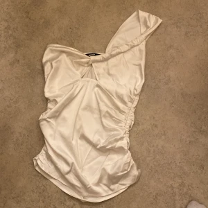 Vit oneshoulder topp från BikBok - Säljer en snygg vit oneshoulder topp från BikBok i storlek XS. Toppen har en elegant drapering och är tillverkad i ett glansigt material som ger en lyxig känsla. Perfekt för festliga tillfällen! ✨