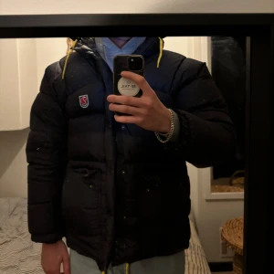 Svart dunjacka från Fjällräven - Säljer en svart dunjacka från Fjällräven i mycket bra skick. Jackan har en pufferstil med långa ärmar och en praktisk dragkedja. Perfekt för kalla vinterdagar med sin varma och quiltade design. Den har också en huva med gula detaljer för extra stilpoäng. Passar perfekt för både vardag och utomhusaktiviteter.