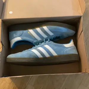 Säljer ett par snygga blå Adidas Spezial sneakers i bra skick med mindre defekter. De har en klassisk design med vita ränder och en mörk sula. Perfekta för både vardag och fest. Skorna är tillverkade i mocka och har snörning för en säker passform. Hör av er ifall ni har några frågor!