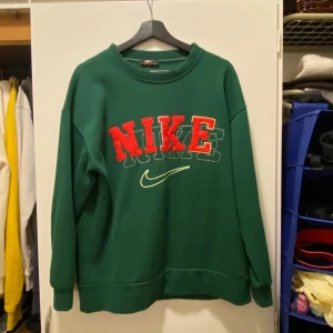 Grön sweatshirt från Nike - Säljer en grön Nike sweatshirt i storlek M. Den har ett stort rött Nike-tryck på framsidan med en cool broderad effekt. Perfekt för höst och vinter med sin mysiga och varma känsla. Passar både till vardags och chilliga dagar hemma. 🟢🔴