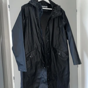 Svart regnjacka från Åhléns - Säljer en svart regnjacka från Åhléns i storlek XS. Jackan har en lång modell med huva och knappar framtill. Perfekt för regniga dagar och håller dig torr och bekväm. Den har långa ärmar och praktiska fickor med knappar.