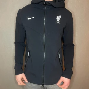 Svart Tech fleece från Nike med LFC-logga - Säljer en svart Nike-tech fleece med LFC-logga. Tech fleecen har dragkedja och huva, perfekt för höst och vår. Den är i mycket bra skick och har en sportig stil. Passar bra för fotbollsfans eller som en casual hoodie.