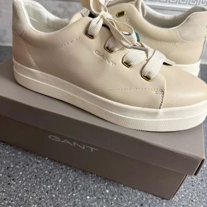 Beige sneakers från GANT - Säljer ett par snygga beige sneakers från GANT i nytt skick. Skorna har en stilren design med snörning och är tillverkade i syntetmaterial. Perfekta för vår och sommar, dessa sneakers är både bekväma och trendiga. Kommer i originalkartong.