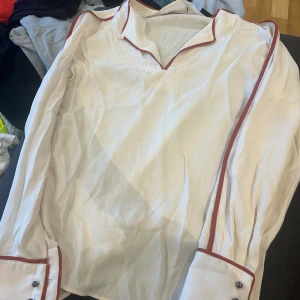 Vit blus från Tiger of Sweden - Säljer en elegant vit blus från Tiger of Sweden. Blusen har långa ärmar och är dekorerad med röda kanter längs kragen, ärmarna och knäppningen. Den har en klassisk krage och knappar vid ärmsluten. Perfekt för både vardag och fest!