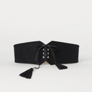 Svart skärp med snörning och tofsar - Säljer ett snyggt svart skärp med snörning och tofsar. Skärpet är brett och har en cool design med guldfärgade öljetter och svarta snören som knyts framtill. Perfekt för att ge din outfit en extra touch! Skärpet är i nyskick och passar de flesta storlekar.