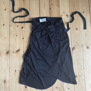 Svart omlottkjol från Maison Margiela - Säljer en svart omlottkjol från Maison Margiela i italiensk storlek 42, vilket är typ 36-38. Kjolen är tillverkad i Italien och har en elegant och enkel design med knytband i midjan. Perfekt för både vardag och fest!