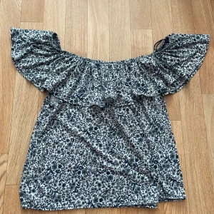 Blommig off-shoulder topp från H&M - Säljer en superfin blommig off-shoulder topp från H&M. Den är vit med ett svart och blått blommönster och har en volangdetalj runt axlarna. 🤗