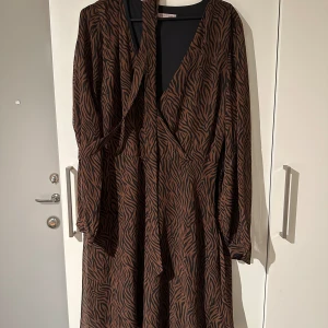 Brun långklänning med zebramönster - Klänning från Anna Field. brun med zebramönster. Klänningen har långa ärmar och en omlott-design framtill. 