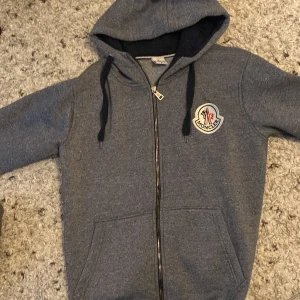Grå hoodie från Moncler byte funkar  - Säljer en snygg grå hoodie från Moncler. Den är i bra skick men dragkedjan är lite sönder så man får dra upp kedjan snabbt. Hoodien har en broderad Moncler-logga på bröstet och är perfekt för höst och vinter. Den är supermjuk och bekväm, perfekt för både vardag och chill.