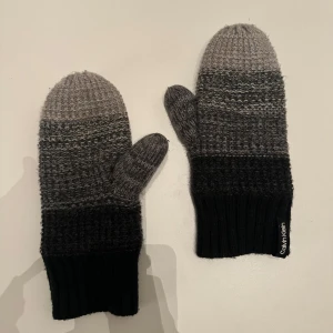 Grå och svarta vantar från Calvin Klein - Säljer ett par snygga och varma vantar från Calvin Klein. De är stickade i olika nyanser av grått och svart, perfekta för kalla vinterdagar. Vantarna har en mjuk insida som håller händerna varma och bekväma. Använda men väldigt bra skick