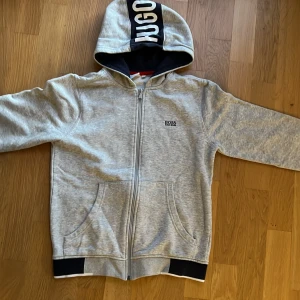 Grå hoodie från Hugo Boss - Säljer en snygg grå hoodie från Hugo Boss i storlek XS. Den har en dragkedja framtill och en stor logga på huvan. Perfekt för en avslappnad stil och passar bra till både höst och vår. Den är i mycket bra skick och redo för nya äventyr!