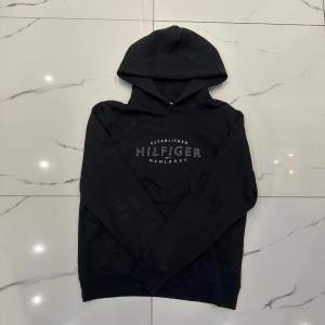 Säljer en snygg svart hoodie från Tommy Hilfiger i storlek M. Den har en klassisk design med Hilfiger-loggan tryckt på framsidan. Perfekt för en casual look och passar till alla tillfällen. Superbekväm och stilren!