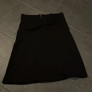 Svart kjol med dragkedja - Snygg svart kjol i klassisk stil med dragkedja bak. Perfekt för både vardag och festliga tillfällen. Kjolen har en enkel och stilren design som gör den lätt att matcha med olika toppar och accessoarer.