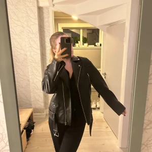 Svart skinnjacka från NA-KD  - Säljer en snygg svart skinnjacka från Nakd i mycket bra skick. Den har en klassisk bikerstil med dragkedjor och knappar som detaljer. Perfekt för höst och vår, och funkar lika bra till fest som till vardags. Jackan har en normal passform och långa ärmar.