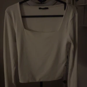 Vit långärmad topp - Säljer en stilren vit långärmad topp i mjukt material. Perfekt för både vardag och festliga tillfällen. Toppen har en enkel design med en fyrkantig halsringning som ger en modern touch.