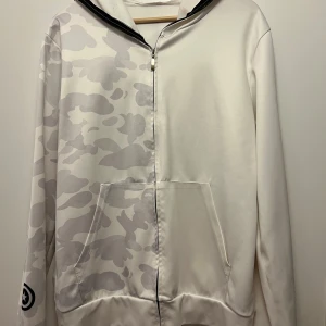 Vit hoodie med kamouflagemönster och hajmotiv - Säljer en cool vit hoodie med grått kamouflagemönster och hajmotiv på huvan. Den har en dragkedja framtill och stora fickor. Perfekt för streetwear-stilen och att sticka ut i mängden!