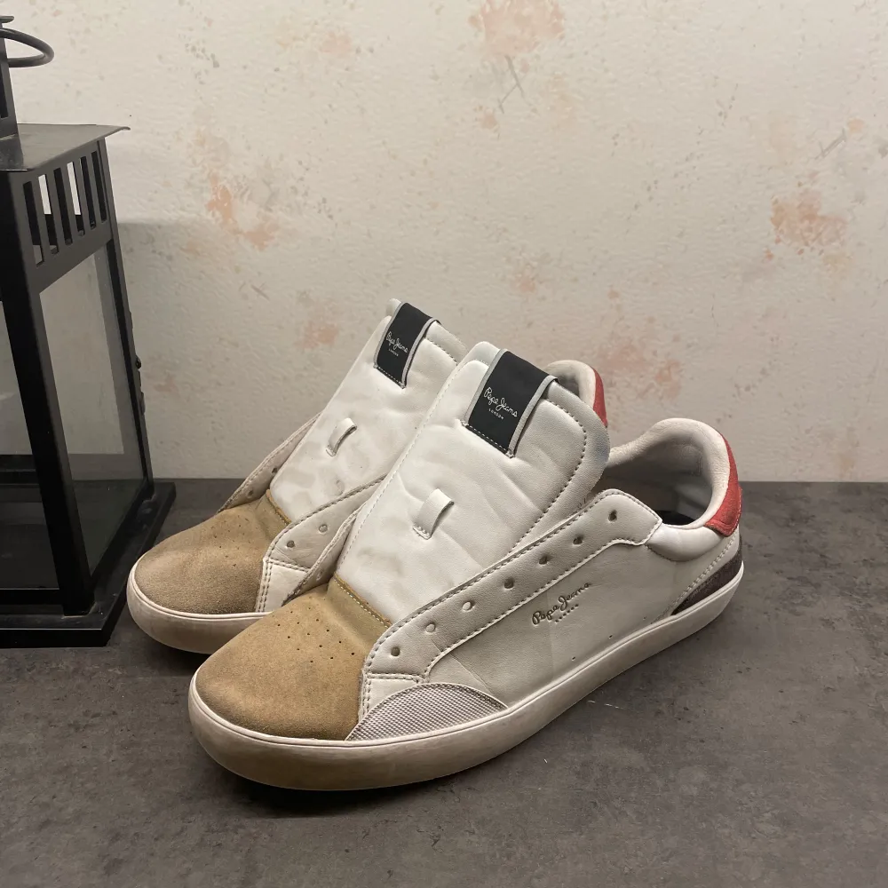 Säljer ett par vita sneakers från pepe jeans bra skick. Skorna har en snygg kombination av vitt och beige med röda detaljer på hälen. De är tillverkade i skinn och mocka, vilket ger en stilren look. Perfekta för både vardag och fest!👌Ny pris 1299. skosnörren sätts självklart på innan.. Skor.