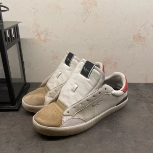 Vita sneakers från Pepe Jeans  - Säljer ett par vita sneakers från pepe jeans bra skick. Skorna har en snygg kombination av vitt och beige med röda detaljer på hälen. De är tillverkade i skinn och mocka, vilket ger en stilren look. Perfekta för både vardag och fest!👌Ny pris 1299. skosnörren sätts självklart på innan.