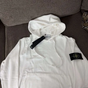 Stone Island hoodie - Säljer en snygg vit hoodie från Stone Island i nytt skick. Den har en klassisk design med en känguruficka och den ikoniska loggan på ärmen. Perfekt för både vardag och chill dagar. Passar till allt och är superbekväm!