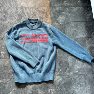 Stickad tröja från J.Lindeberg - Säljer en snygg blå tröja från J.Lindeberg med röd text och mönster på bröstet. Tröjan har en klassisk rund hals och långa ärmar med ribbade muddar. Perfekt för en stilren och avslappnad look. Passar både till vardags och lite finare tillfällen. Storlek M men passar S. Skicket är bra då tröjan är i hög kvalitet!