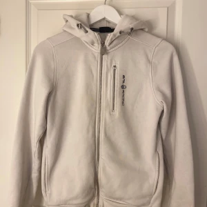 Vit fleecejacka från Sail Racing - Säljer en vit fleecejfrån Sail Racing i mycket bra skick. Den har en dragkedja framtill och en praktisk huva med justerbara snören. Ordinarie pris runt 1000 kr