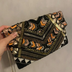 Färgglad fest kuvertväska med pärlor - Säljer en supercool kuvertväska med ett färgglatt mönster i svart, guld och orange. Väskan är dekorerad med massor av pärlor och har en kedja som axelrem. Perfekt för en fest eller en utekväll när du vill sticka ut! 👜✨