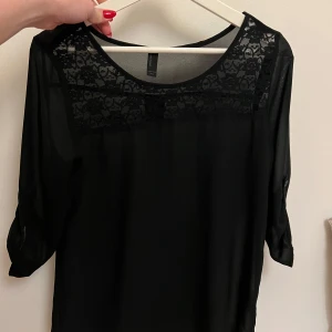 Svart blus med spetsdetaljer - Snygg svart blus i medium storlek med spetsdetaljer vid halsringningen. Blusen har trekvartsärmar och en luftig passform, perfekt för både vardag och fest. Den är gjord i ett lätt och bekvämt material som ger en elegant look.
