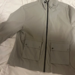 Grå vindjacka med dragkedja - Snygg grå vindjacka med dragkedja och två stora fickor framtill. Jackan har en stilren design och är perfekt för blåsiga dagar. Den har en hög krage och knappdetaljer på fickorna.