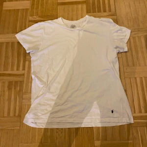 Vit t-shirt från Polo Ralph Lauren - Säljer en klassisk vit t-shirt från Polo Ralph Lauren i 100% bomull. T-shirten har en liten broderad logotyp i svart på framsidan. Perfekt för en stilren och avslappnad look.