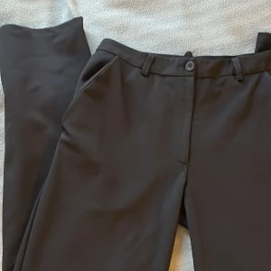 Svarta kostymbyxor - Snygga svarta kostymbyxor med medelhög midja och raka ben. Byxorna har en klassisk design med knappar och dragkedja fram. De är insydda i midjan men det går att sprätta. Midjemåttet är 36cm rakt över och innerbenslängden är 83cm. 