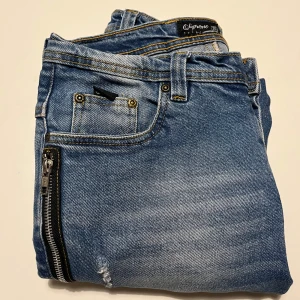 Blå jeans med dragkedja och slitningar - Snygga blå jeans från Supreme med coola slitningar och en dragkedja på sidan. Perfekta för en trendig look. De har en klassisk femficksdesign och är tillverkade i ett bekvämt denimtyg.