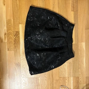 Svart kjol från Gina Tricot - Snygg svart kjol från Gina Tricot med glansig yta och subtilt mönster. Perfekt för att ge en elegant touch till din outfit. Kjolen har en kort längd och en bekväm passform.