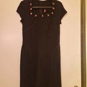 Svart klänning med dekorativa detaljer - Elegant svart klänning med korta ärmar och dekorativa detaljer vid halsringningen. Perfekt för en stilren 38 look.