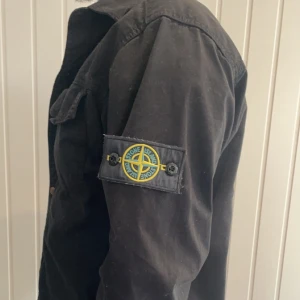 Svart overshirt från Stone Island - Snygg svart overshirt från Stone Island med klassisk logga på ärmen. Skjortan har knappar framtill och två bröstfickor. Perfekt för lager-på-lager-stil. Skriv för mer bilder och pris går att diskutera