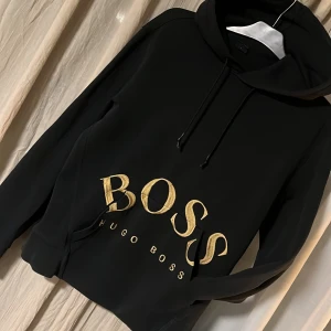 Svart hoodie från Hugo Boss - Snygg svart hoodie från Hugo Boss med stort guldtryck på framsidan. Den har justerbar huva och långa ärmar. 