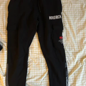 Svarta cargobyxor från Hoodrich - Snygga svarta cargobyxor från Hoodrich med flera fickor och coola detaljer. Byxorna har en avslappnad passform och är perfekta för en streetwear-look. De har en justerbar midja med snörning och tryck på sidan.