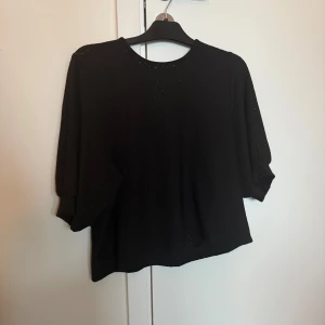 Svart blus med trekvartsärm - Elegant svart blus med trekvartsärm och rund halsringning. Blusen har en knappdetalj i nacken och är perfekt för en stilren look.