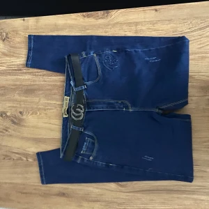 Blå jeans med bälte - Snygga blå jeans med ett svart bälte och silverfärgat spänne. Jeansen har en klassisk design med fem fickor och subtila slitningar för en trendig look.